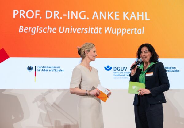 Die Moderatorin lächelt die Laudatorin an, die ein Mikrofon in der Hand hält. Beide stehen auf einer Bühne in festlicher Kleidung.