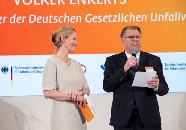 Die Moderatorin lächelt den Laudator an, der ein Mikrofon in der Hand hält. Beide stehen auf einer Bühne in festlicher Kleidung.